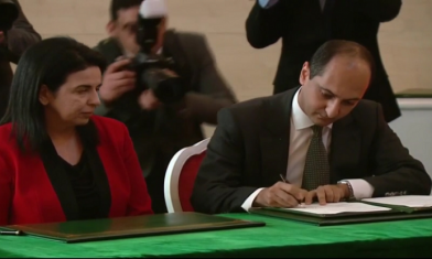 Signature d’une convention entre un représentant masculin en costume et une représentante féminine en veste rouge, devant un parterre de photographes.